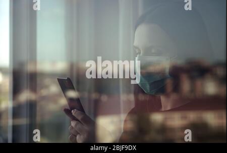 Woman wearing mask and using smartphone behind closed window Stock Photo