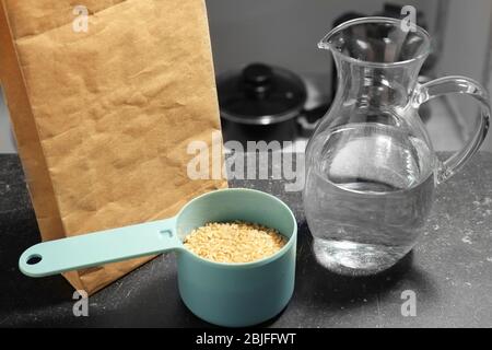 Measure scoop with brown rice and jug with water on kitchen table Stock Photo