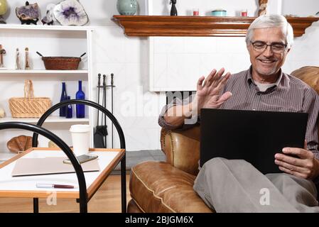 Senior man waving at his laptop screen as he video chats with family while under stay at home restrictions due to the COVID-19 pandemic. Stock Photo