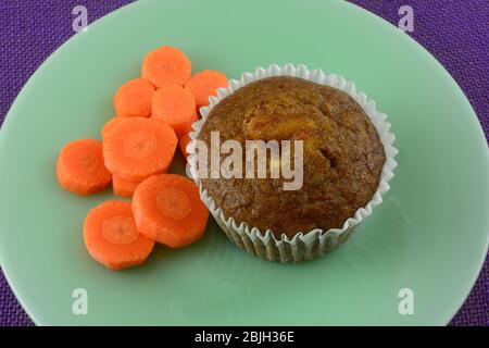 Baked carrot muffin in white paper muffin cup with carrot slices on green breakfast plate on purple tablecloth Stock Photo