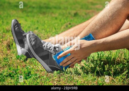 Young man applying cold compress to leg while sitting on grass outdoors, closeup Stock Photo