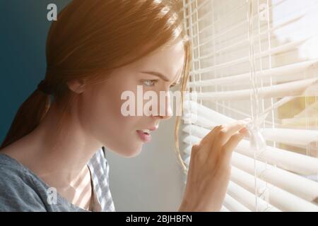 Beautiful young girl separating slats of blinds and looking through window Stock Photo