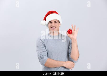 Handsome man in Christmas hat holding shiny bauble on light background Stock Photo