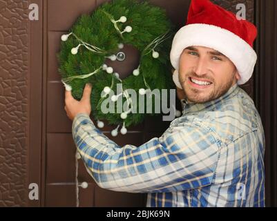 Man in Santa hat hanging Christmas wreath on door Stock Photo