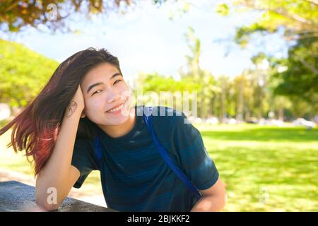 Smiling biracial Asian Caucasian teen girl or young woman sitting at table outdoors in park on sunny day, leaning head on hand and looking at camera, Stock Photo