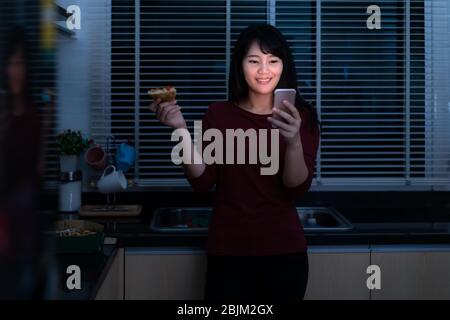 Asian woman virtual happy hour meeting and eating delivery pizza from the box online with friend or taking photo using mobile phone camera in kitchen Stock Photo