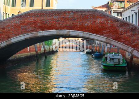 Old red brick walking bridge over the water canals of venice, Italy at dawn with boats tied to posts Stock Photo