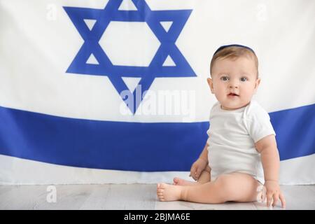 Cute baby in kippah sitting on floor against big flag of Israel Stock Photo