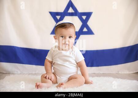 Cute baby in kippah sitting on floor against big flag of Israel Stock Photo