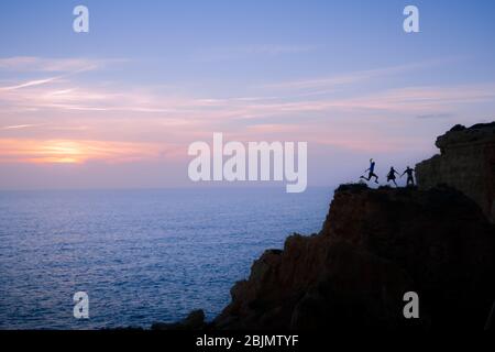A group of 3 friends on vacation jumps at the same time for a picture taken on timer, making up a fun backlight image on top of the cliffs. Stock Photo