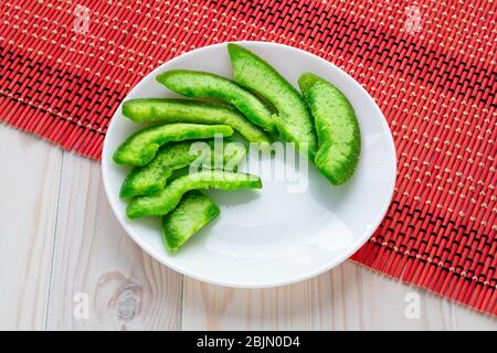 Green slices of succade of pomelo on white plate on wooden light background and bright red wicker dining mat. Candied fruit, confectionery top view Stock Photo