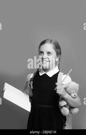 Girl with braids and happy face. Pupil holds blue book, marker and teddy bear. Kid in school uniform isolated on green background. Study and back to school concept. Stock Photo