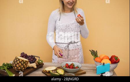 Young woman cutting strawberries fr while streaming online for webinar lesson - Influencer girl making smoothie doing video masterclass with phone cam Stock Photo