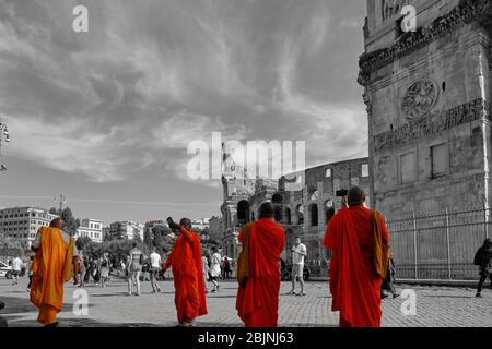 Group of Buddhist monks in orange robes walking through a city taking a selfie in front of the coliseum in Rome, Italy and landmark as people walk by Stock Photo