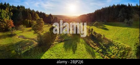 Aerial landscape panorama after sunrise: gorgeous scenery with the sun in the blue sky, trees on green meadows casting long shadows, surrounded by for Stock Photo