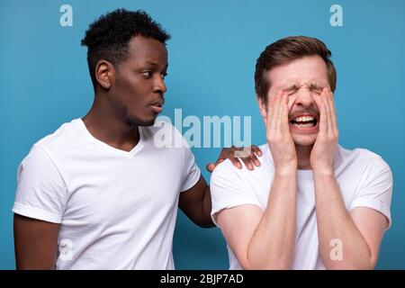 African american man calms down his colleague or friend, who has troubles, miserable facial expressions, keeps hands on his shoulders, stands closely Stock Photo