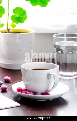 A white cup of coffee with multi-colored sweets stands on a wooden table, next to a stack of books and a notebook against Stock Photo