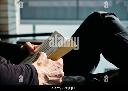 closeup of a young caucasian man, in casual wear, reading a book sitting in an outdoors armchair on the balcony Stock Photo