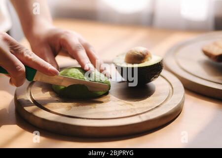 Close up of woman slicing avocado on wooden board at home. Vegetarian food concept Stock Photo