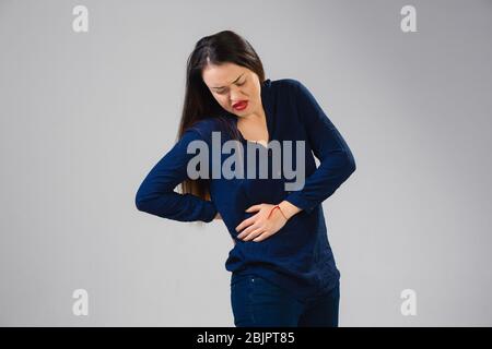 Young caucasian woman suffers from back pain, feels sick, ill and weakness isolted on gray studio background. Symptoms of desease or unhealthy lifestyle. Sickness, ilness, sad and upset. Copyspace. Stock Photo