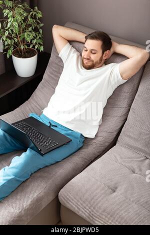 Home work place. Smiling happy relaxed man freelancer working from home using laptop. Cozy home office on sofa. Coronavirus covid-19 social distancing Stock Photo
