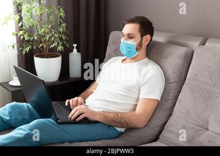 Young caucasian man working from home wearing protective mask using laptop and internet. Cozy Home office, workplace on sofa during coronavirus Stock Photo