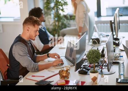 young designer typing on computer keyboard at workplace in the open space office with colleagues in the background. business agency Stock Photo