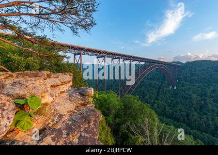 The large steel arch bridge of the New River Gorge spans the canyon below a blue sky viewed from the rock ledge of a climbing area. Stock Photo