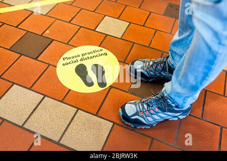 Yellow warning sign to maintain social distance distancing during covid-19 coronavirus outbreak with footsteps in store and legs of person in USA Stock Photo