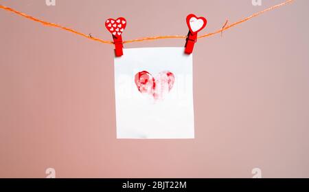 a piece of paper with a patterned heart of fingerprints of red clothespins with a heart on a rope. Valentine's Day love. pink background. copy space. Stock Photo