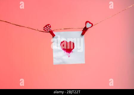 a piece of paper with a patterned heart of fingerprints of red clothespins with a heart on a rope. Valentine's Day love. pink background. copy space. Stock Photo