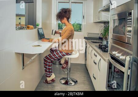 Side view of a black woman working at home during Coronavirus quarantine.  Conference call in the kitchen wearing pajamas and jacket. Stock Photo