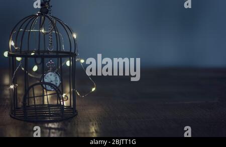 Conceptual photo of imprisonment during Coronavirus confinement.  A clock imprisoned in a bird cage on a wooden table. Selective focus on the clock. Stock Photo