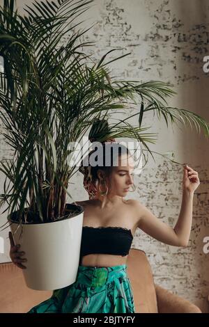 A tanned girl in summer outfit holds large homemade flower in her hands. She touches a leaf of the plant with her hand.  Stock Photo