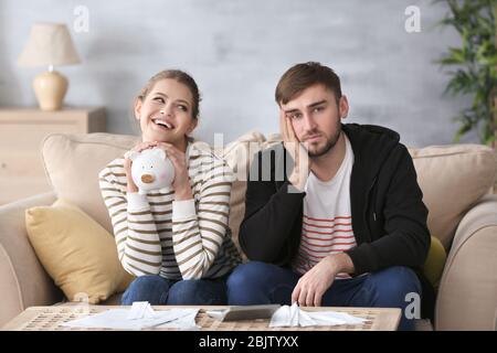 Happy woman holding piggy bank and sad man with bills at home. Money problems in relationship Stock Photo