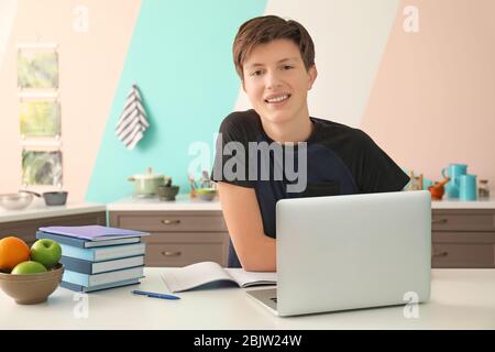 Cute teenager boy using laptop while doing homework in kitchen Stock Photo