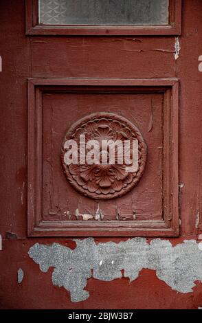 Architectural fretwork element of red painted wooden door with flaking paint. Sculptural rosette in square frame of door panel of classic building in Stock Photo