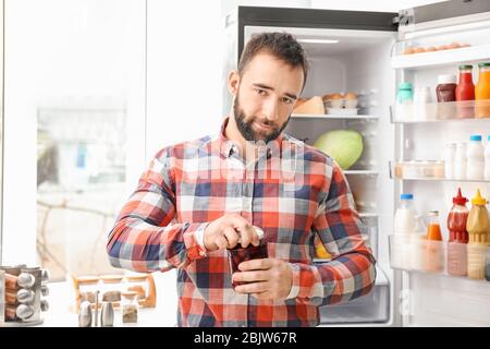 Handsome man opening jar near refrigerator in kitchen Stock Photo