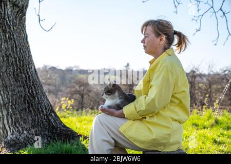 Female in casual clothes sitting near a big tree, holding grey cat on her hands and looking into the distance Stock Photo
