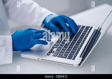 Doctor or scientist in white coat and blue gloves is writing on a laptop in his clinic or laboratory office. Biochemistry and biotechnology concept. Stock Photo
