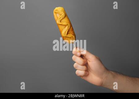 Woman holding tasty corn dog with mustard on gray background Stock Photo