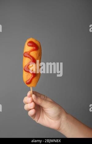 Woman holding tasty corn dog with ketchup on gray background Stock Photo