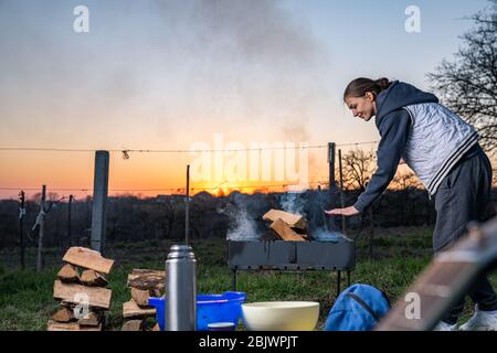 Girl warms up near open fire. Woman holds hands above flame to toast herself. Burning woods logs and charcoal are in barbecue grill Stock Photo