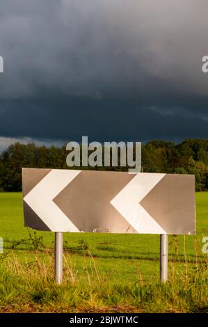 A chevron roadsign warns of a sharp bend on a country road, with dark storm clouds behind. Stock Photo