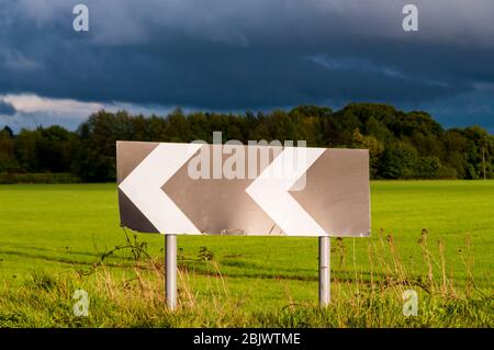 A chevron roadsign warns of a sharp bend on a country road, with dark storm clouds behind. Stock Photo