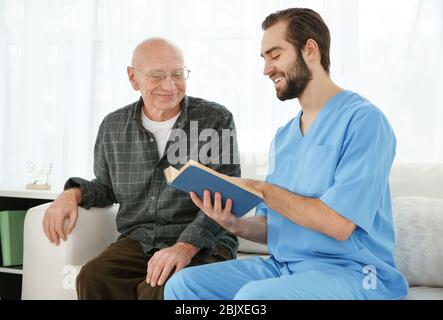 Young caregiver reading book to senior man at home Stock Photo