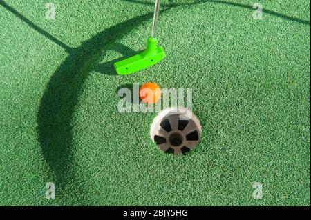 Green mini golf club puts and orange ball towards a hole on green turf Stock Photo