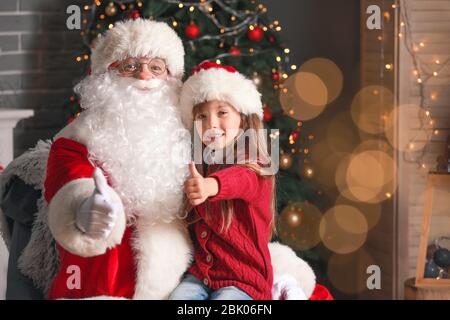 Santa Claus and little girl showing thumb-up in room decorated for Christmas Stock Photo