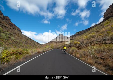 An asphalt road passing through the incredible nature of the mountains in Gran Canaria. A cyclist traveling to the Rocky Volcanic Hills. Grand Canary Stock Photo