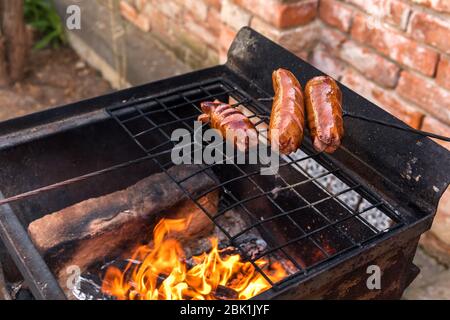 Sausage stabbed on an iron fork and toasted over an fire. Old rusty metal grill. Burning logs in a barbecue grill. Stock Photo